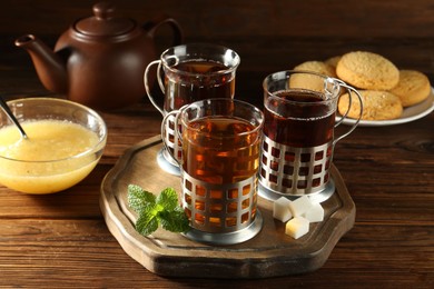 Glasses of hot tea in holders, sugar cubes, mint, honey and cookies on wooden table