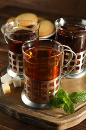 Glasses of hot tea in holders, sugar cubes and mint on wooden table, closeup