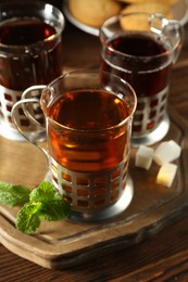 Photo of Glasses of hot tea in holders, sugar cubes and mint on wooden table, closeup