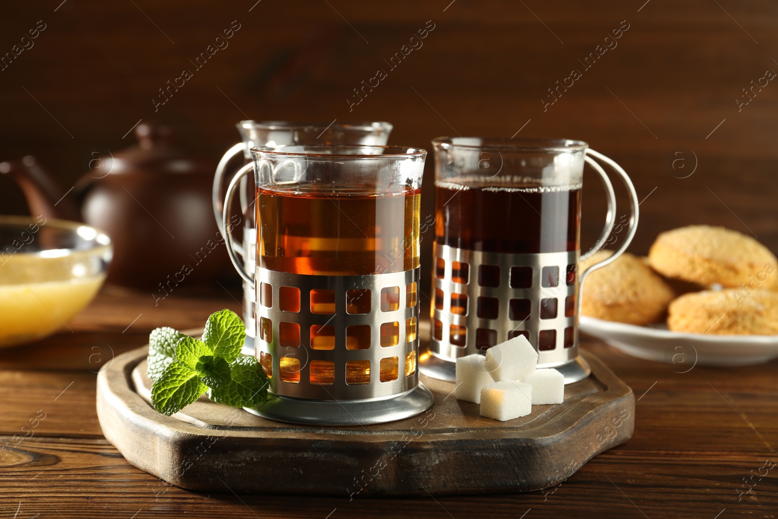 Photo of Glasses of hot tea in holders, sugar cubes, mint, honey and cookies on wooden table, closeup