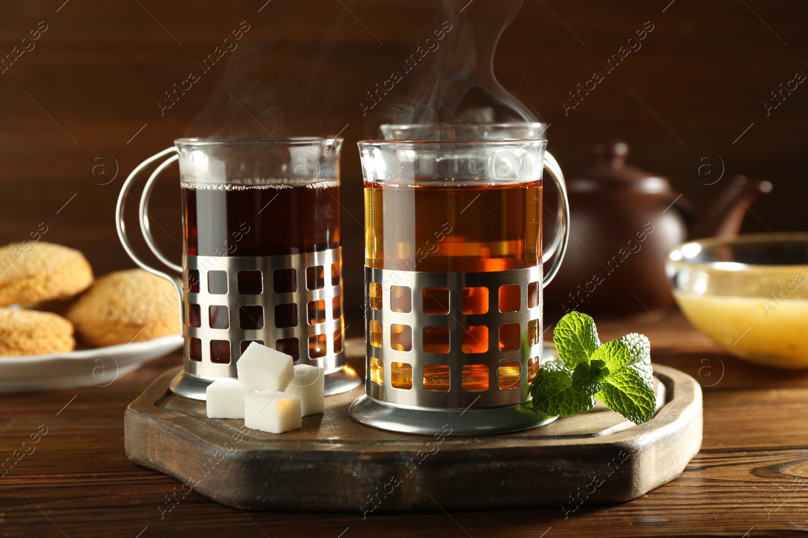 Photo of Glasses of hot tea in holders, sugar cubes, mint, honey and cookies on wooden table, closeup