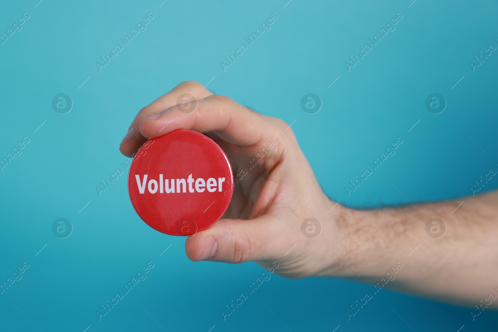 Photo of Man holding button badge with word Volunteer on light blue background, closeup