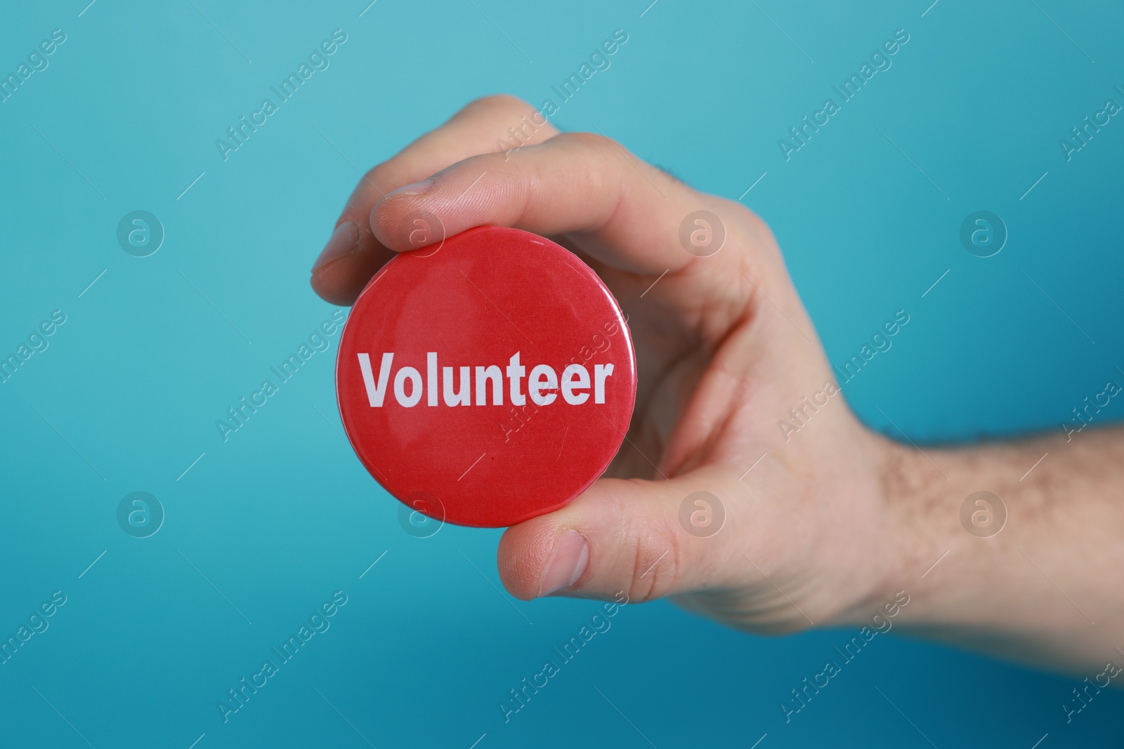 Photo of Man holding button badge with word Volunteer on light blue background, closeup