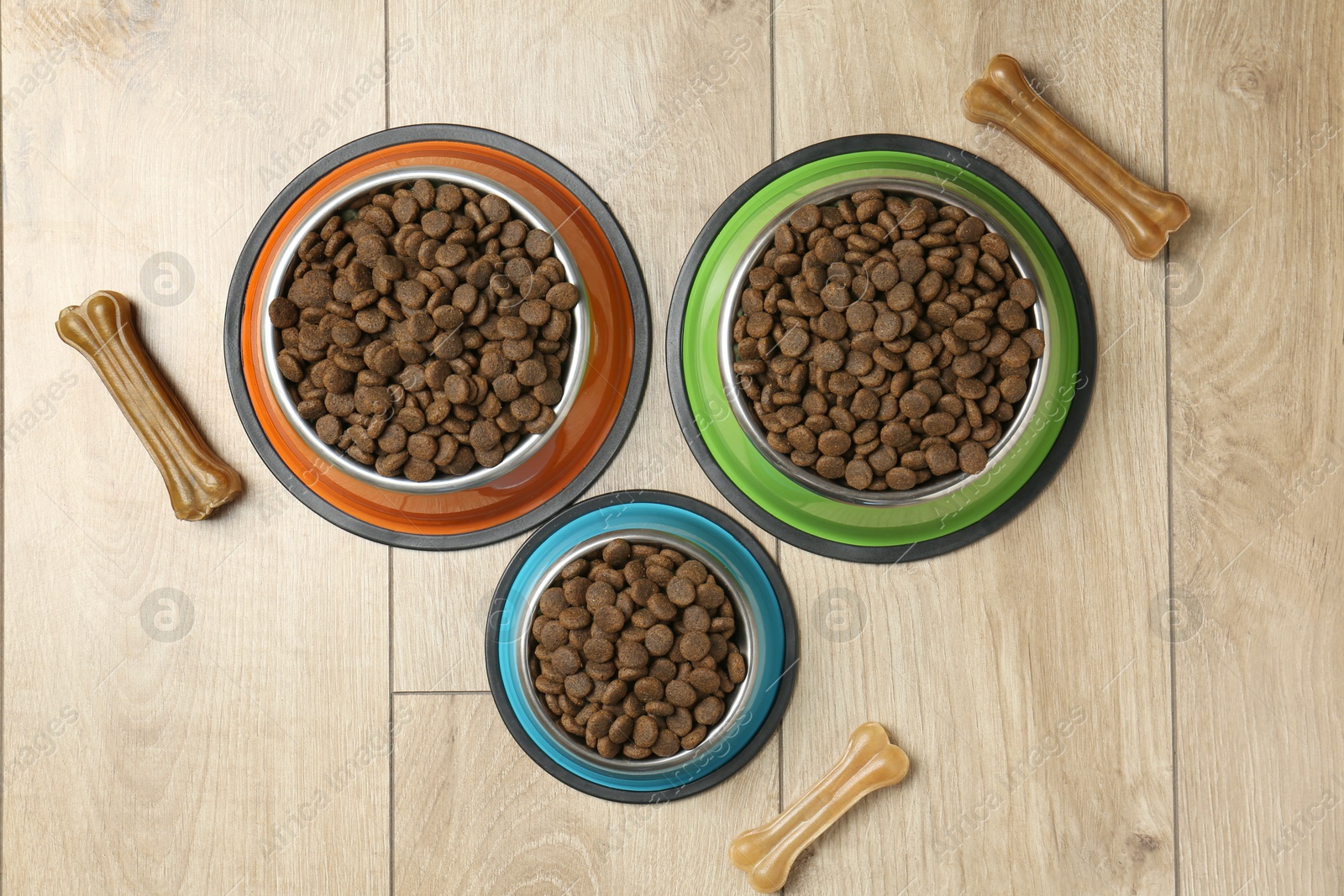 Photo of Dry pet food in feeding bowls and chew bones on wooden floor, flat lay