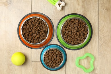 Photo of Dry pet food in feeding bowls and toys on wooden floor, flat lay