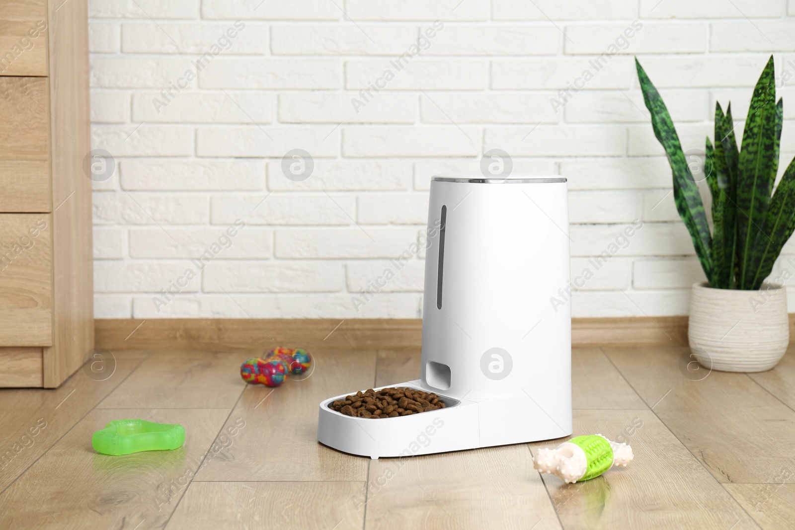 Photo of Dry pet food in automatic feeder and toys on floor indoors