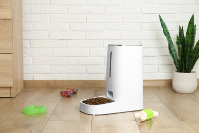 Photo of Dry pet food in automatic feeder and toys on floor indoors