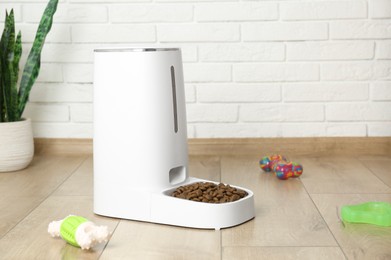 Photo of Dry pet food in automatic feeder and toys on floor indoors