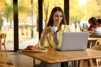 Photo of Woman with coffee and croissant working on laptop at table in cafe