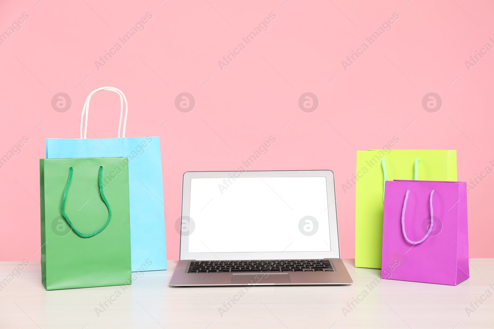 Photo of Internet shopping. Laptop and colorful paper bags on table against pink background