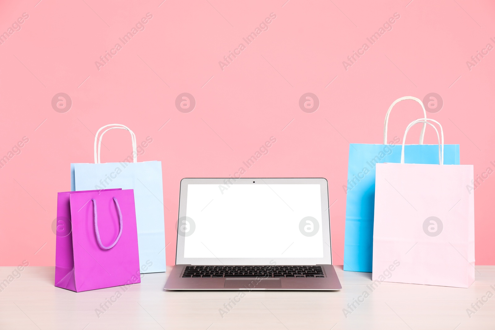 Photo of Internet shopping. Laptop and colorful paper bags on table against pink background