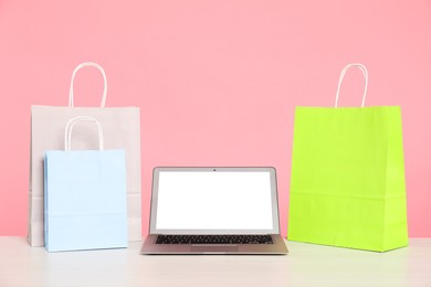 Photo of Internet shopping. Laptop and colorful paper bags on table against pink background