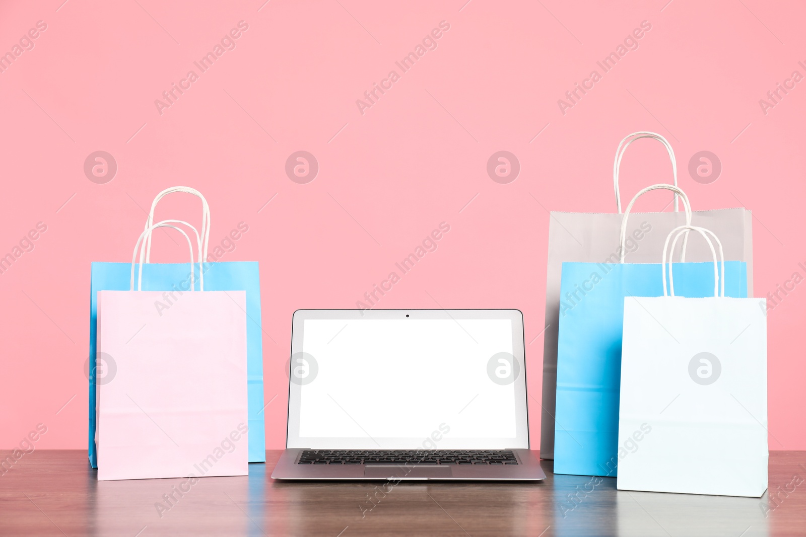 Photo of Internet shopping. Laptop and colorful paper bags on wooden table against pink background