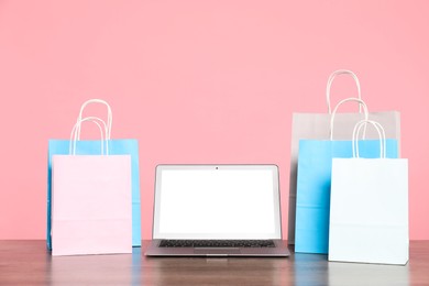 Photo of Internet shopping. Laptop and colorful paper bags on wooden table against pink background