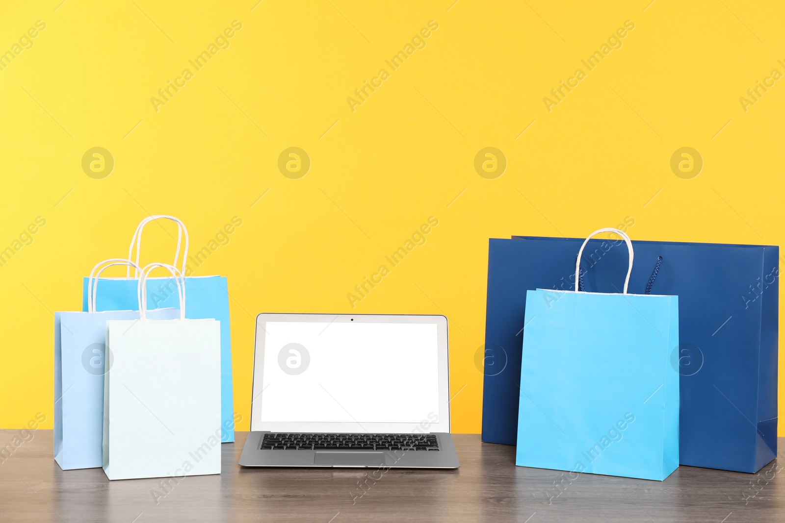 Photo of Internet shopping. Laptop and colorful paper bags on wooden table against yellow background