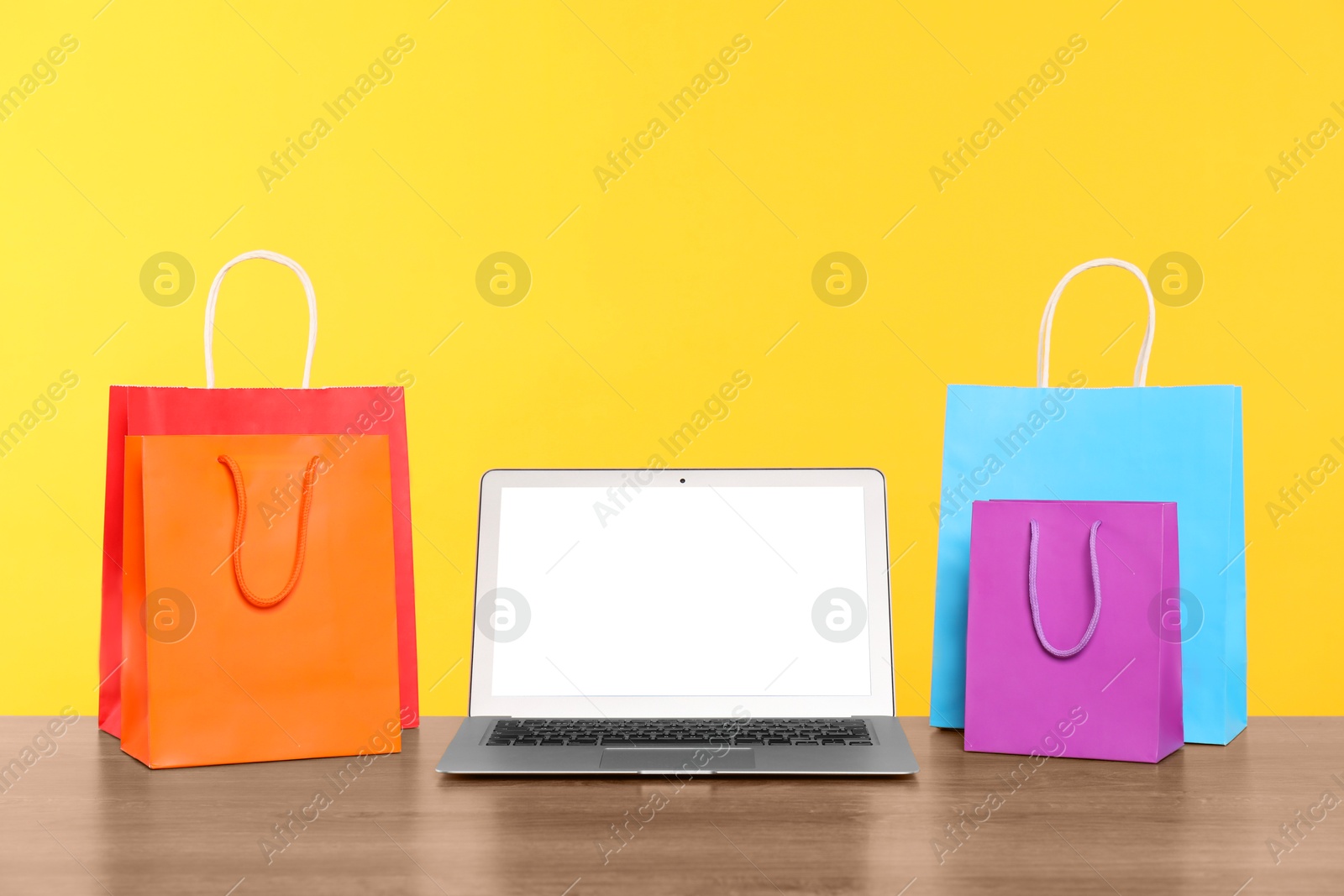 Photo of Internet shopping. Laptop and colorful paper bags on wooden table against yellow background