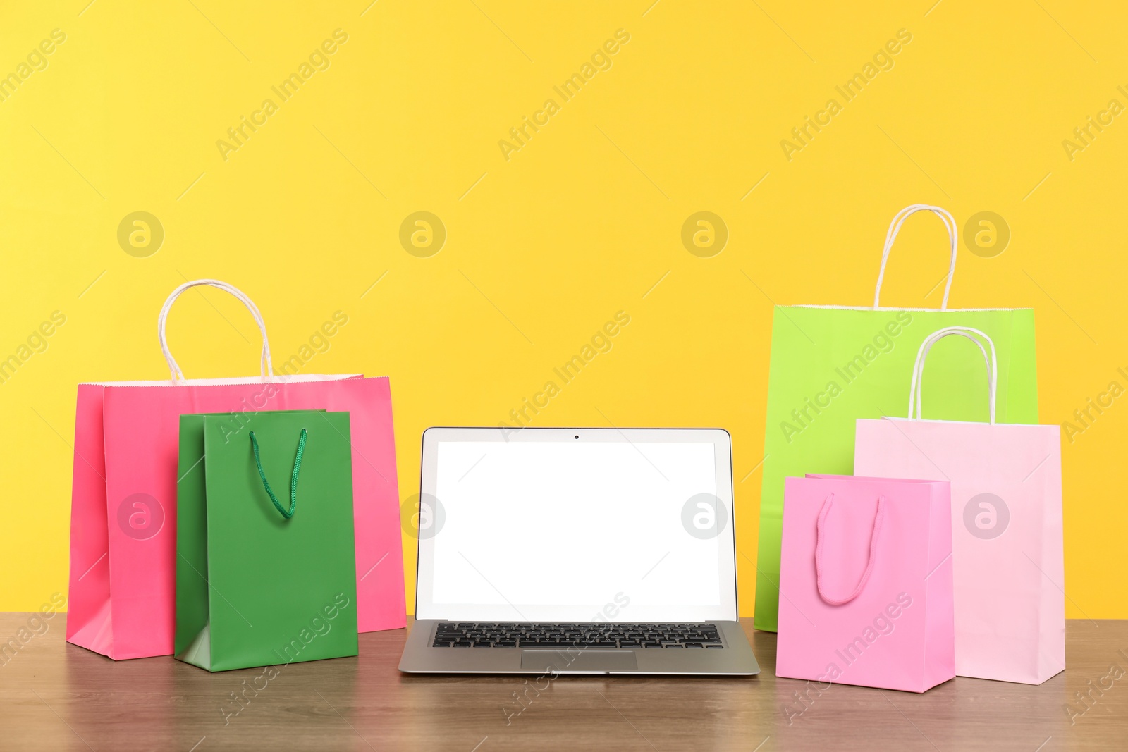 Photo of Internet shopping. Laptop and colorful paper bags on wooden table against yellow background