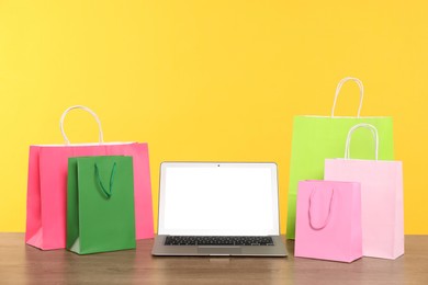 Photo of Internet shopping. Laptop and colorful paper bags on wooden table against yellow background