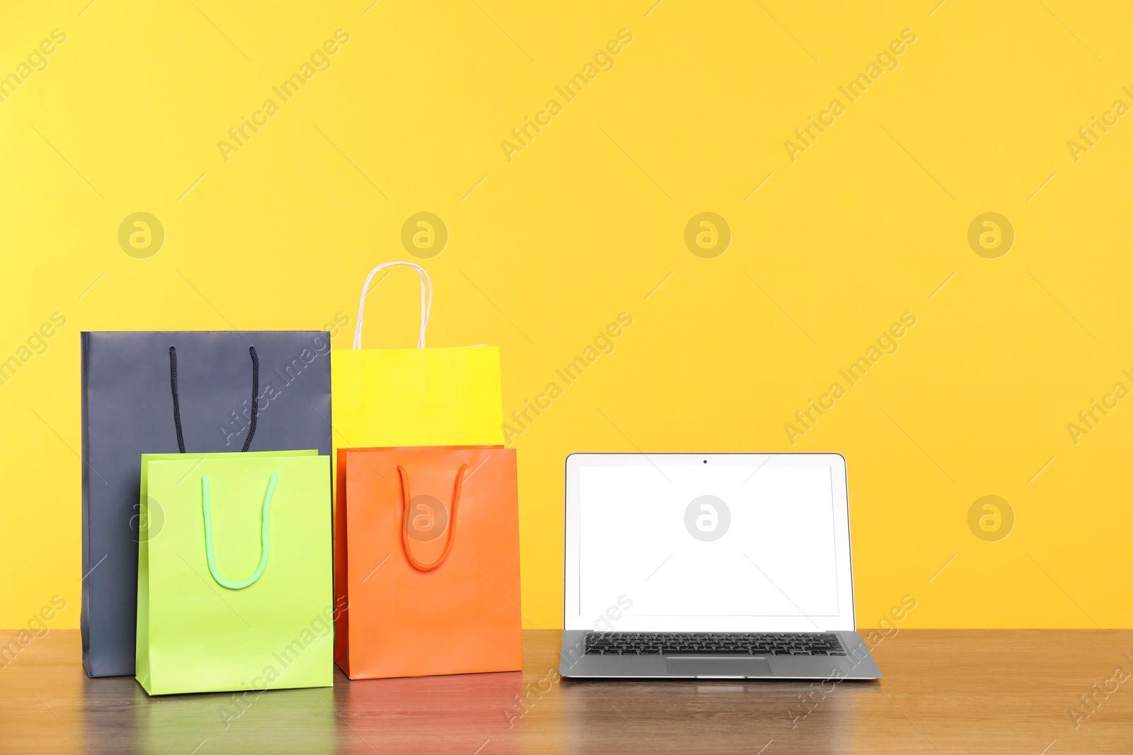 Photo of Internet shopping. Laptop and colorful paper bags on wooden table against yellow background