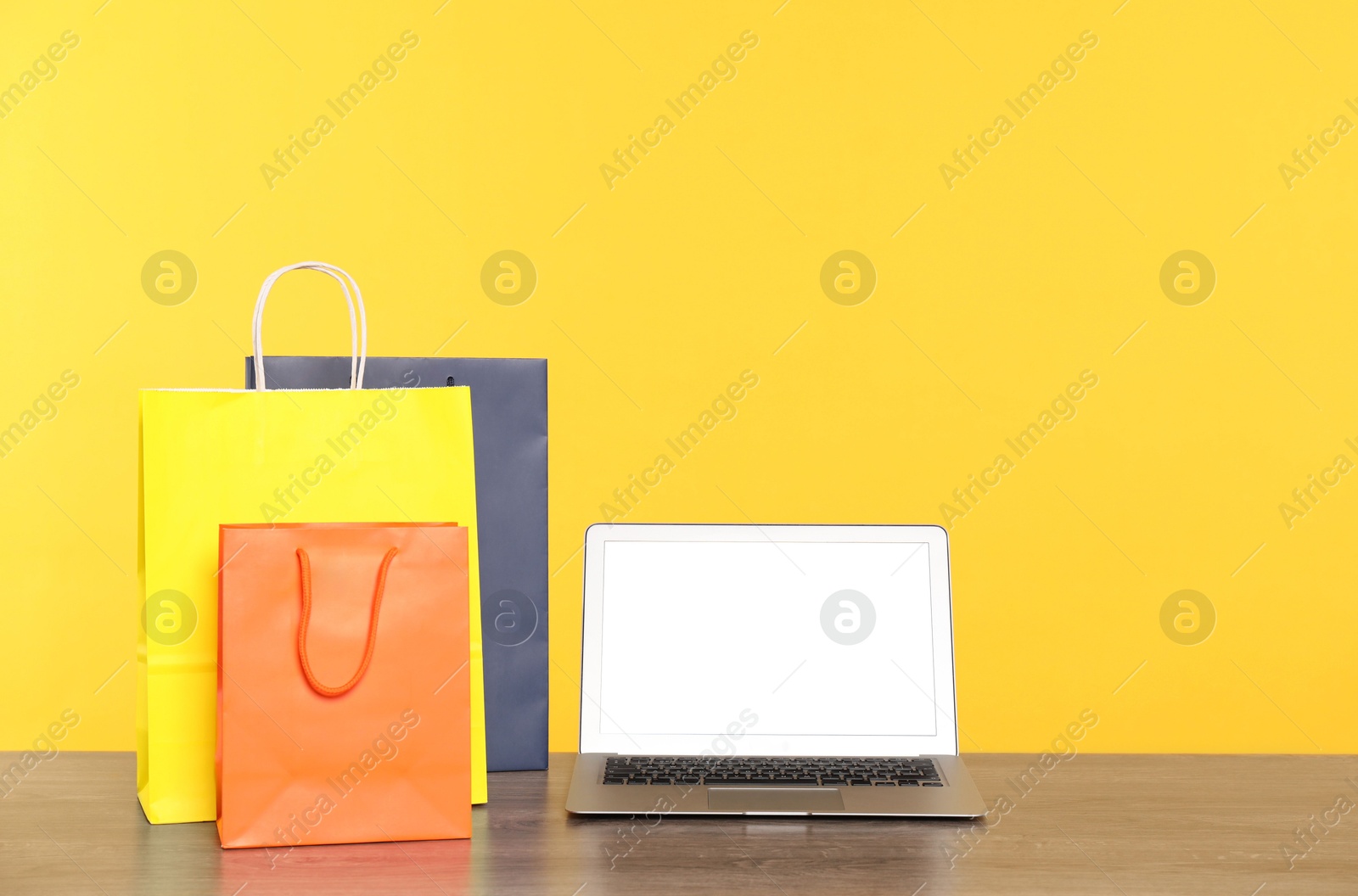 Photo of Internet shopping. Laptop and colorful paper bags on wooden table against yellow background