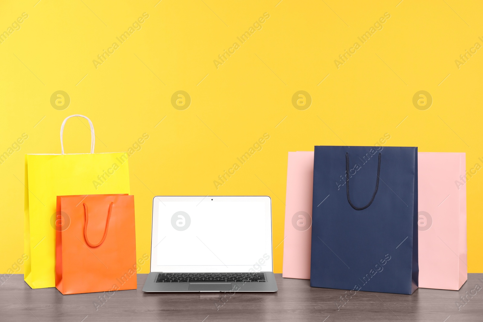 Photo of Internet shopping. Laptop and colorful paper bags on wooden table against yellow background