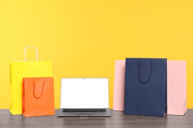 Photo of Internet shopping. Laptop and colorful paper bags on wooden table against yellow background