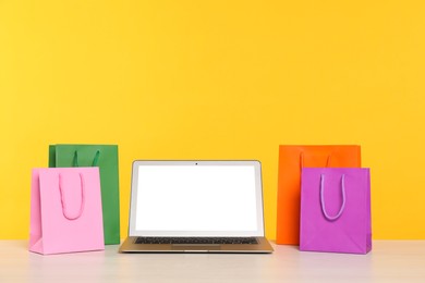 Photo of Internet shopping. Laptop and colorful paper bags on table against yellow background