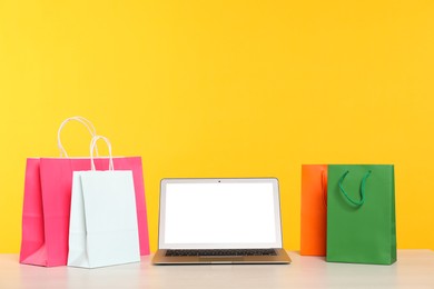 Photo of Internet shopping. Laptop and colorful paper bags on table against yellow background