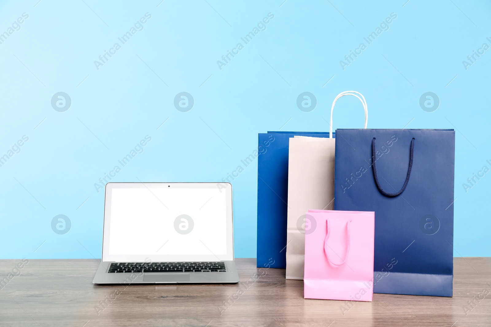 Photo of Internet shopping. Laptop and colorful paper bags on wooden table against light blue background