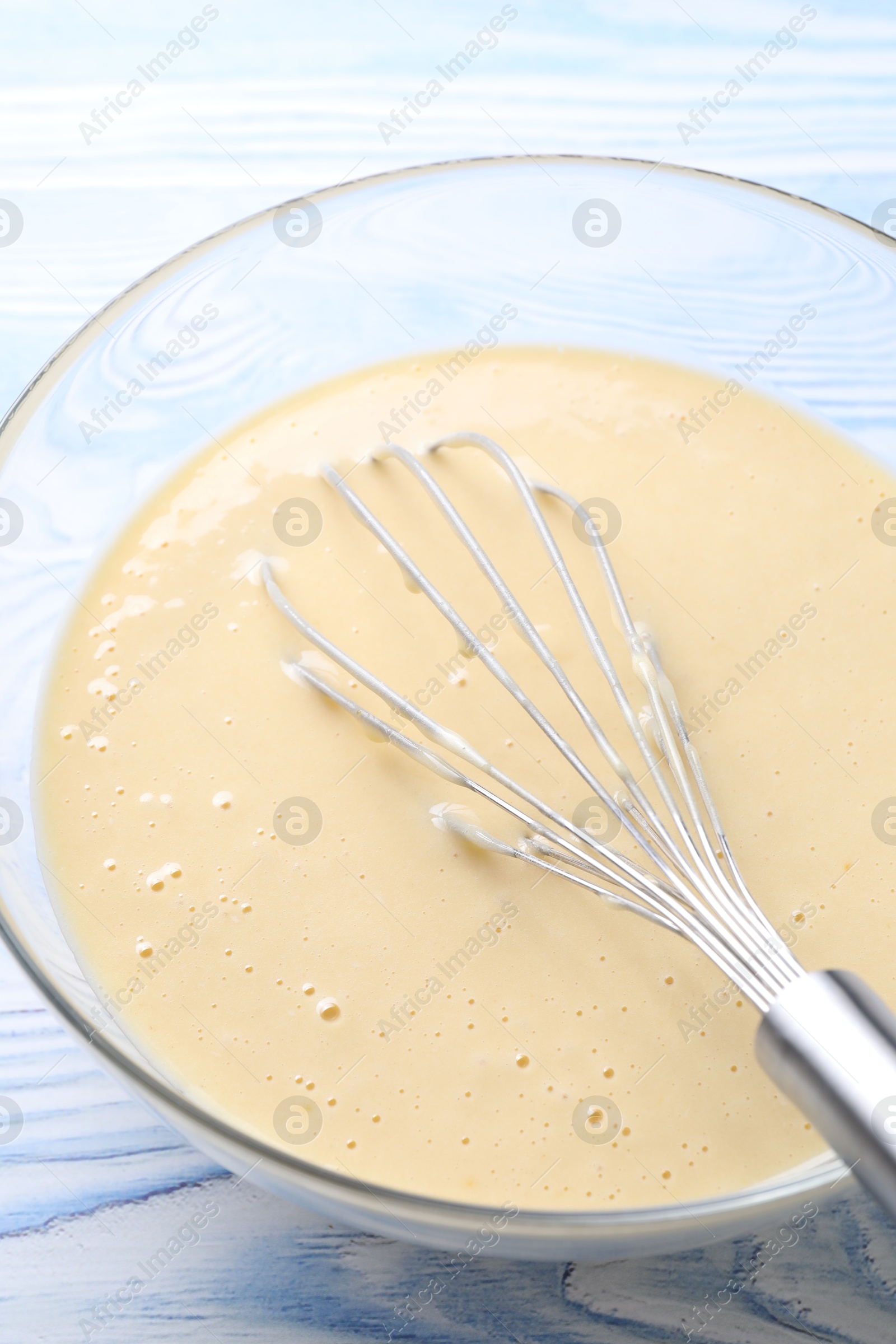 Photo of Whisk and bowl of dough on light blue wooden table, closeup