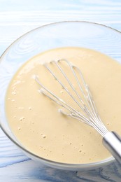 Photo of Whisk and bowl of dough on light blue wooden table, closeup