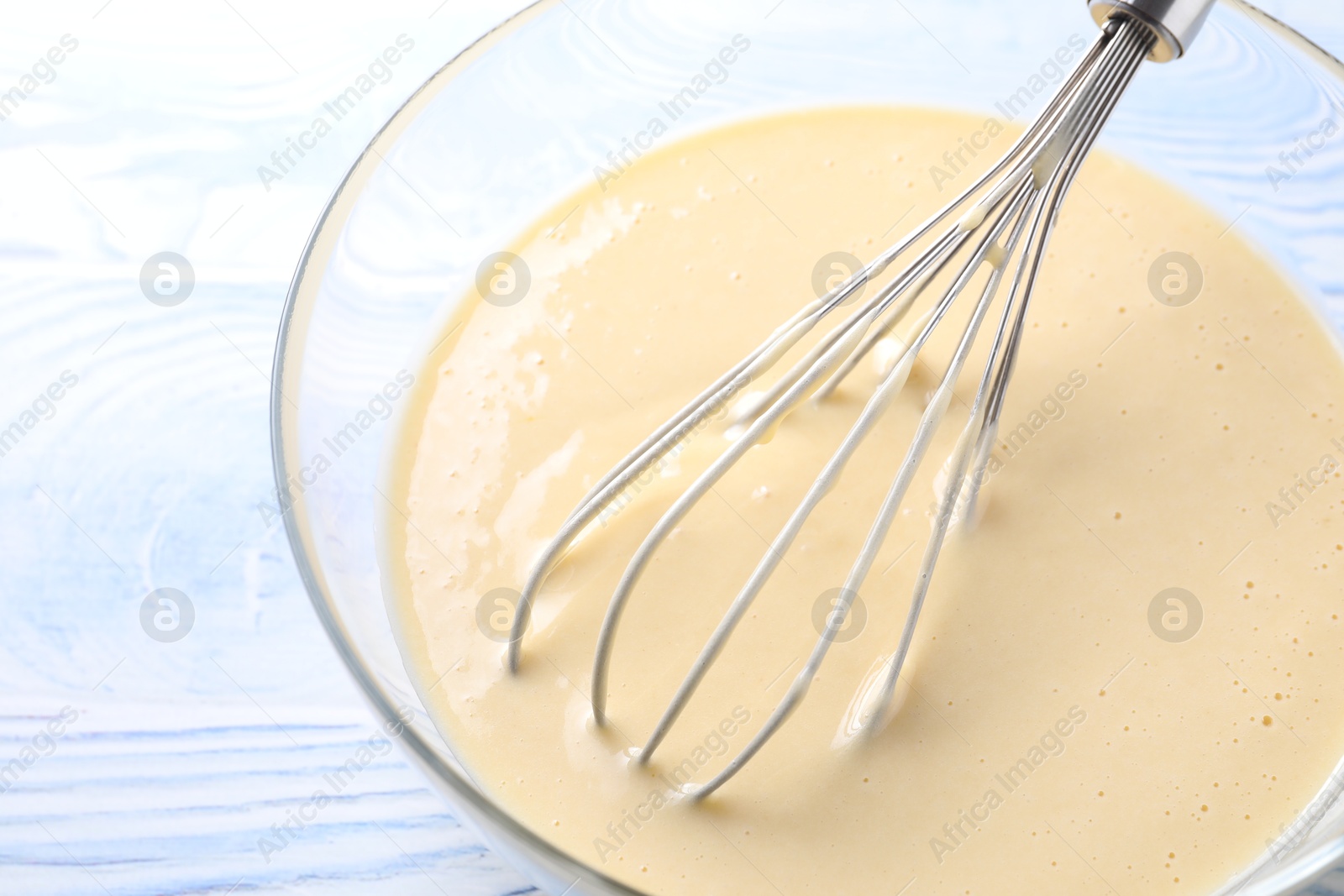 Photo of Whisk and bowl of dough on light blue wooden table, closeup