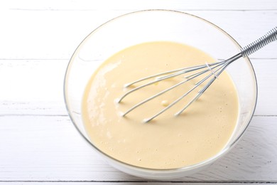 Photo of Whisk and bowl of dough on white wooden table, closeup