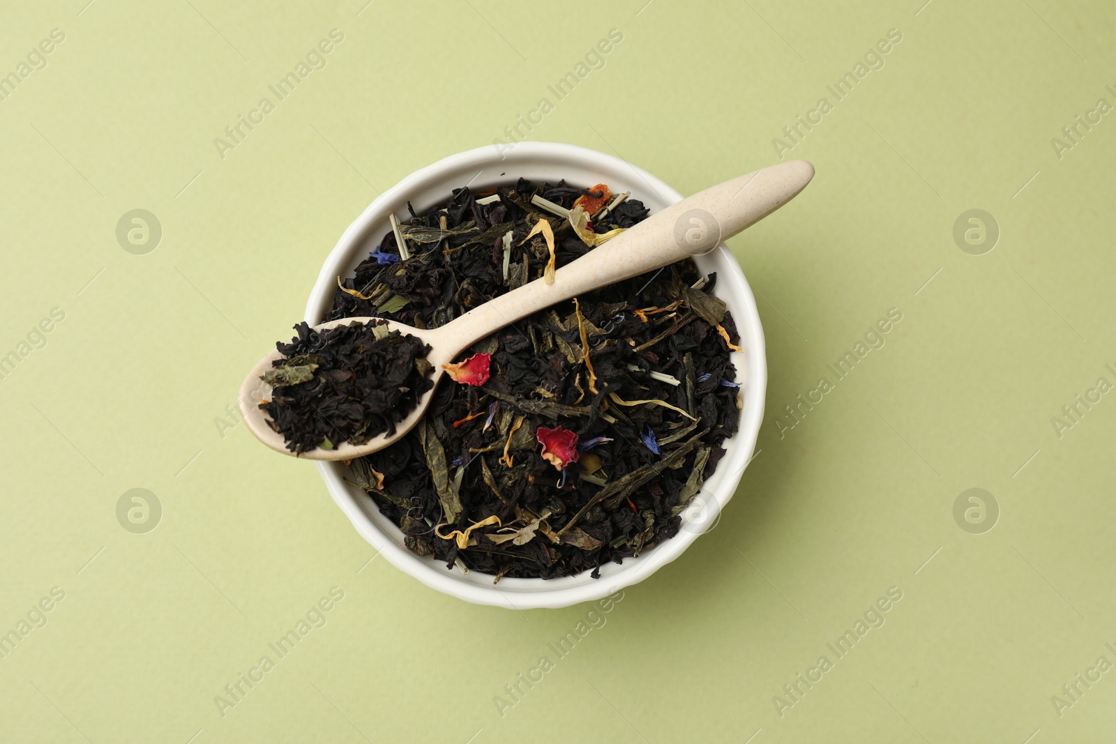 Photo of Dried tea leaves in bowl and spoon on olive background, top view