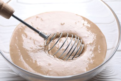 Photo of Whisk and bowl of dough on white wooden table, closeup