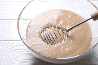 Photo of Whisk and bowl of dough on white wooden table, closeup