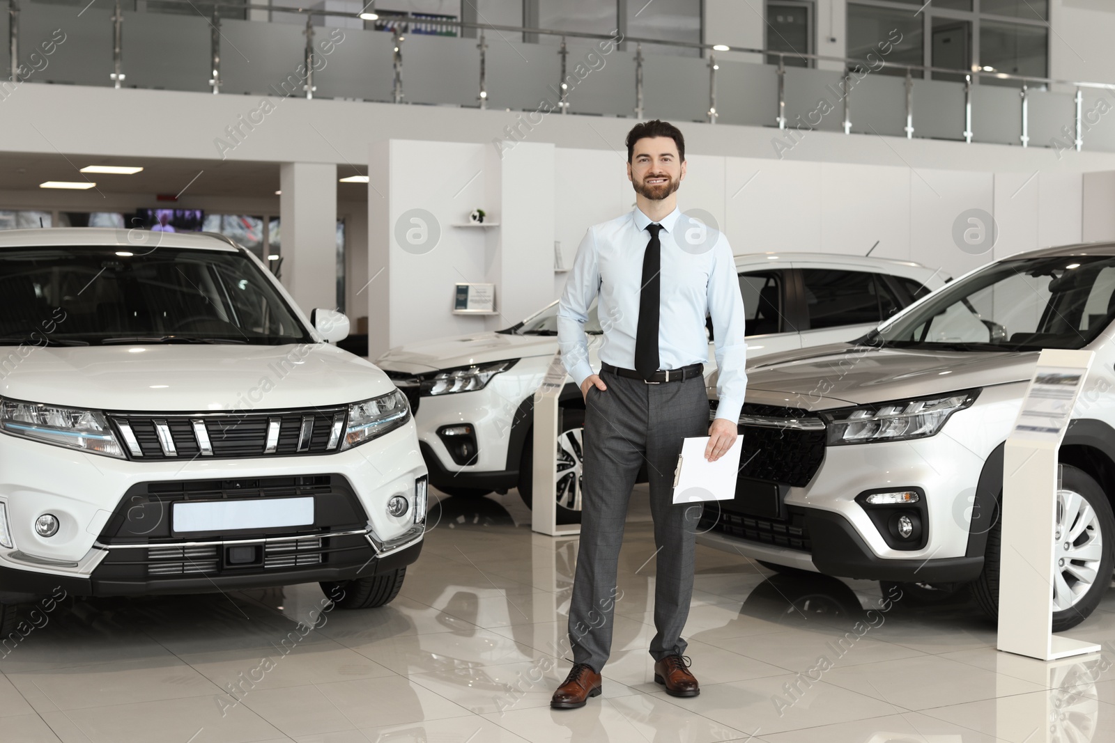 Photo of Happy salesman with clipboard near new cars in salon