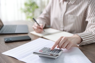 Photo of Budget. Woman with calculator, paperwork and laptop at wooden desk indoors, closeup