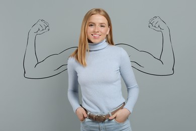 Beautiful young woman with drawing of strong arms behind her on grey background