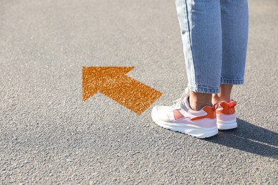 Image of Woman standing near orange arrow on asphalt road, closeup. Concepts of choice, decision, moving forward