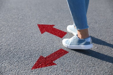 Image of Woman walking on asphalt road with red arrows pointing in different directions, closeup. Concepts of choice, decision, alternative way