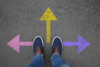 Image of Man standing near colorful arrows on asphalt road, closeup. Concepts of choice, decision, different way, alternative