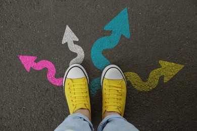 Image of Woman standing near colorful arrows pointing in different directions on asphalt road, closeup. Concepts of choice, decision, alternative way