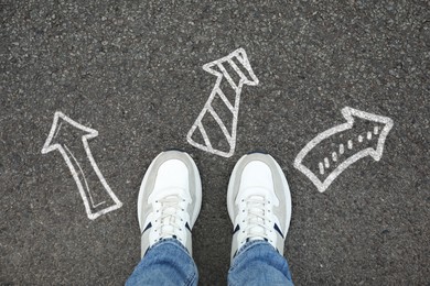 Image of Man standing near white arrows pointing in different directions on asphalt road, closeup. Concepts of choice, decision, alternative way