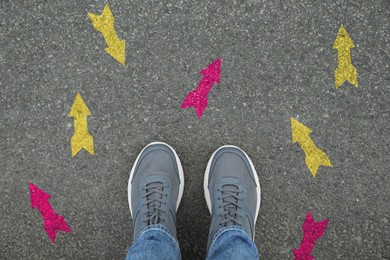 Image of Man standing near yellow and pink arrows pointing in different directions on asphalt road, closeup. Concepts of choice, decision, alternative way