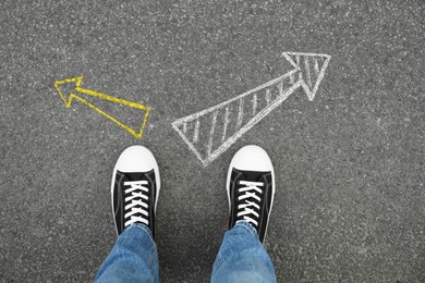 Image of Man standing near white and yellow arrows pointing in different directions on asphalt road, closeup. Concepts of choice, decision, alternative way