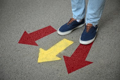 Image of Man standing near red and yellow arrows pointing in different directions on asphalt road, closeup. Concepts of choice, decision, alternative way