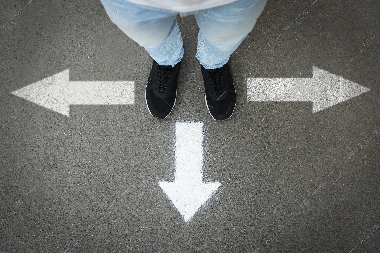 Image of Man standing near white arrows pointing in different directions on asphalt road, closeup. Concepts of choice, decision, alternative way