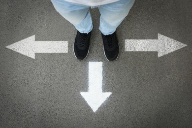 Image of Man standing near white arrows pointing in different directions on asphalt road, closeup. Concepts of choice, decision, alternative way