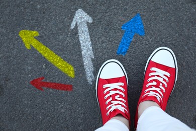 Image of Woman standing near colorful arrows pointing in different directions on asphalt road, closeup. Concepts of choice, decision, alternative way